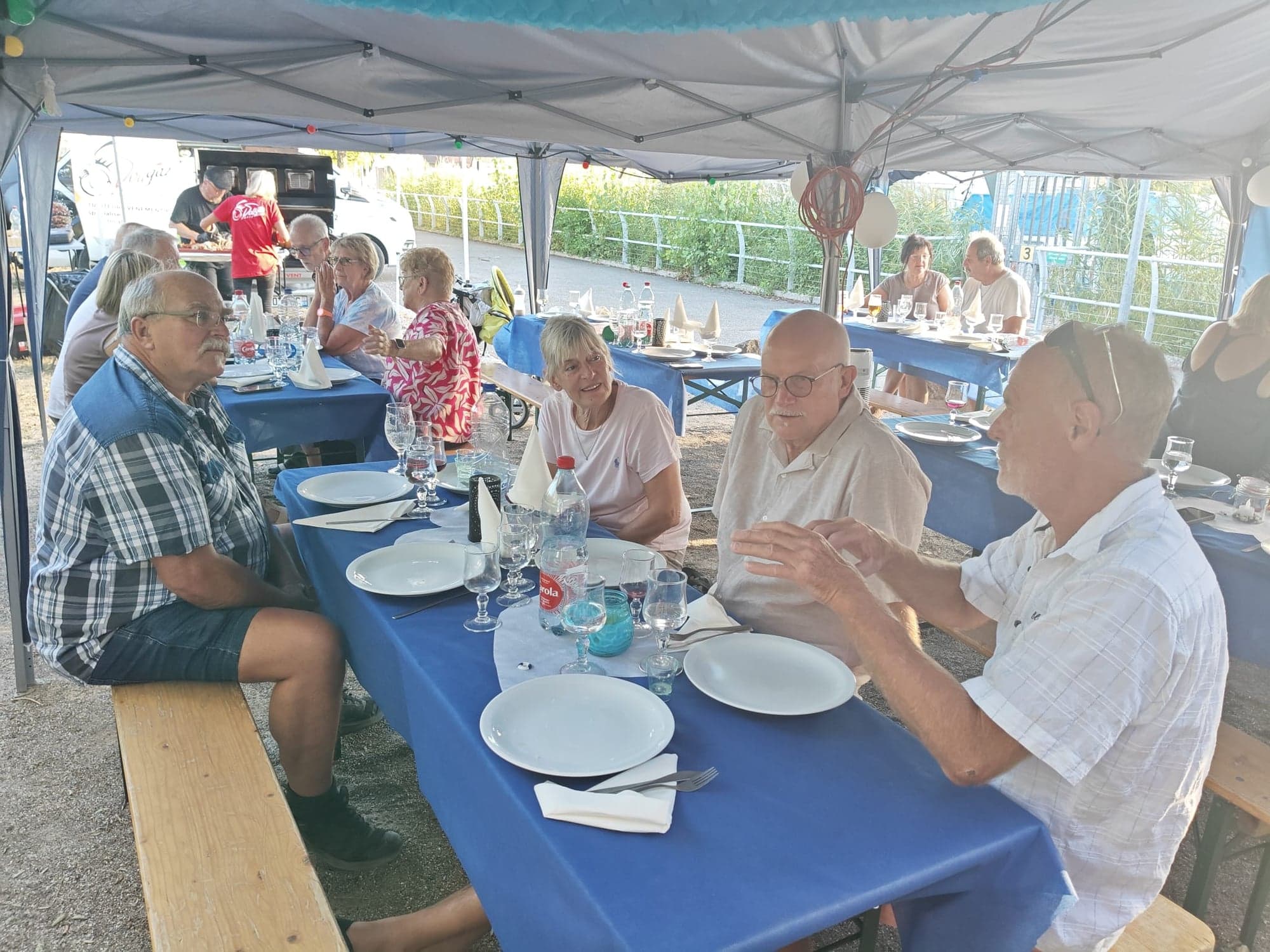 Repas du club sous barnum : membres du Nautic Club Kembs attablés autour de longues tables nappées de bleu, sur des bancs de brasserie, le stand du traiteur en arrière-plan